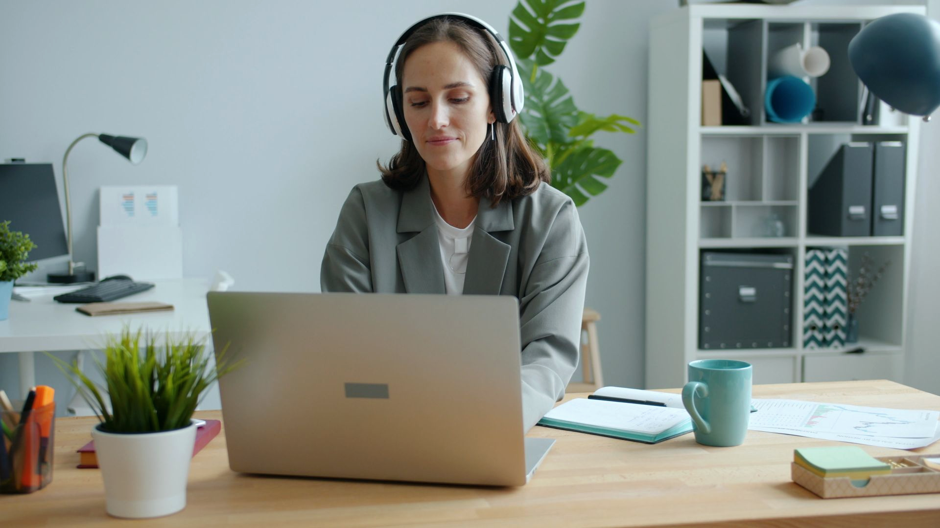 A woman with headphones sits at a home desk looking at her laptop after a call, with a teal mug, amber sticky notes, and a real room behind her.