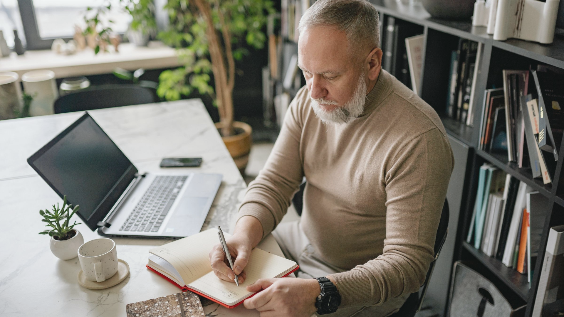 A gray-bearded man in a beige sweater sits at a white marble desk in a home office, writing in a red planner beside an open laptop, a phone, and a coffee cup, with bookshelves and a potted tree behind him.