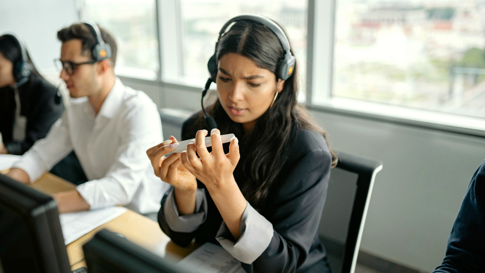 Sales professional wearing a headset at a shared call desk, looking at a phone mid-block, with a colleague on a headset beside her in a bright office bullpen.