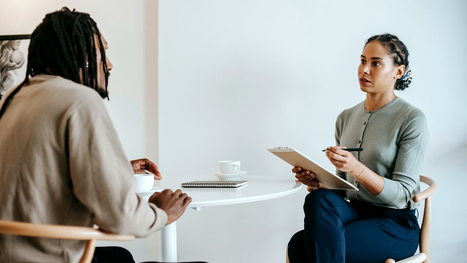 Coach and client in a real one-on-one consultation at a small table with a clipboard and notebook in a calm indoor coaching room.