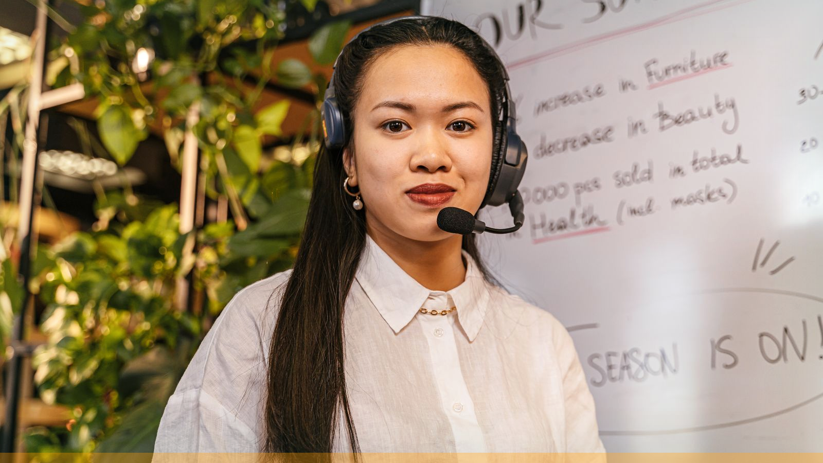 Professional woman with headset and boom microphone in a real call-heavy workspace; whiteboard and plants behind her; photographic stock for live unsupervised speaking pressure.