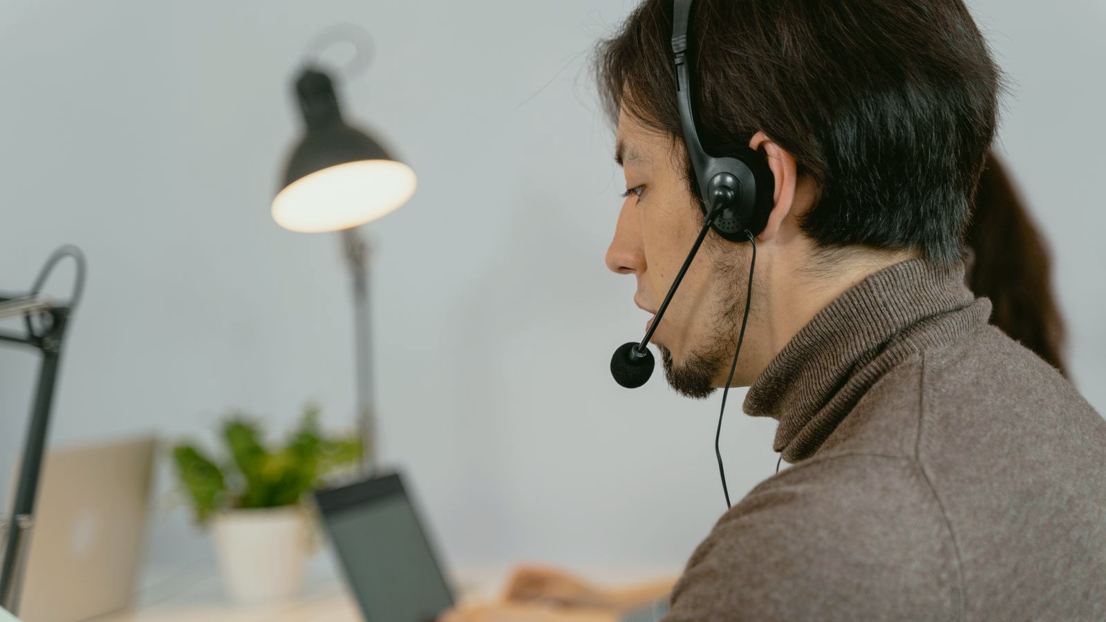 Contact-center professional with a headset mid-call at a desk, human face visible in profile, warm desk lamp and a colleague blurred behind; real collections or call-center environment.