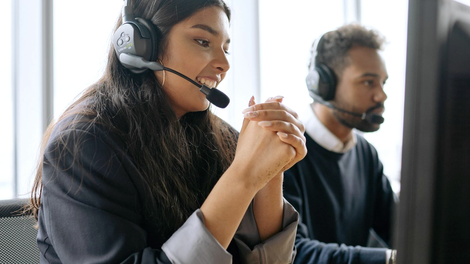 Contact-center agent wearing a headset at a shared desk, smiling while focused on a call, with a colleague in a headset working at the next station behind her.