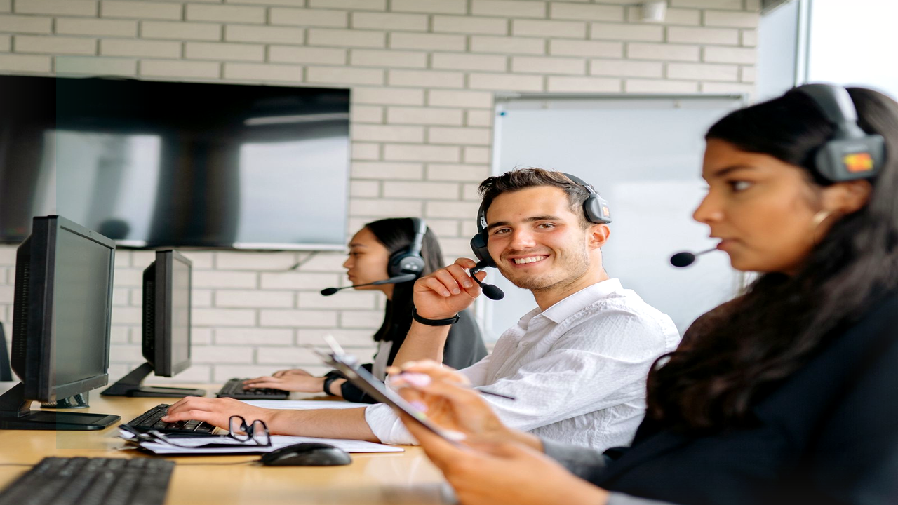 Contact-center agent in a white shirt wearing a headset and smiling toward the camera at a shared desk with monitors, clipboard, and other headset agents working nearby on a collections-style floor.