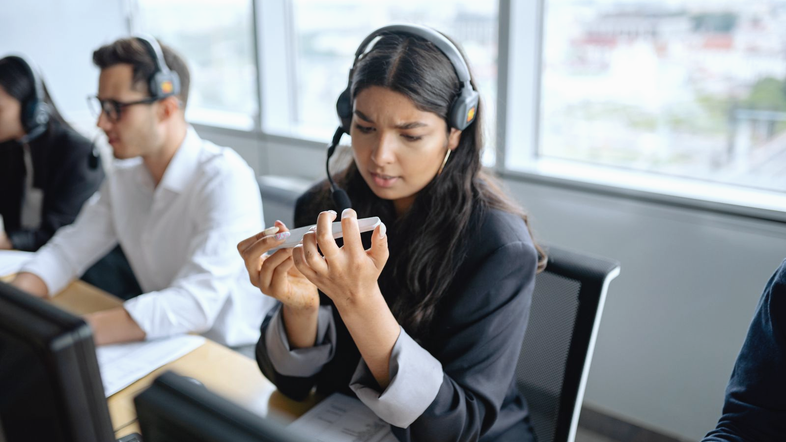 Contact-center agent wearing a headset at a shared desk, brow furrowed in concentration mid-call, with colleagues in headsets working at neighboring stations.