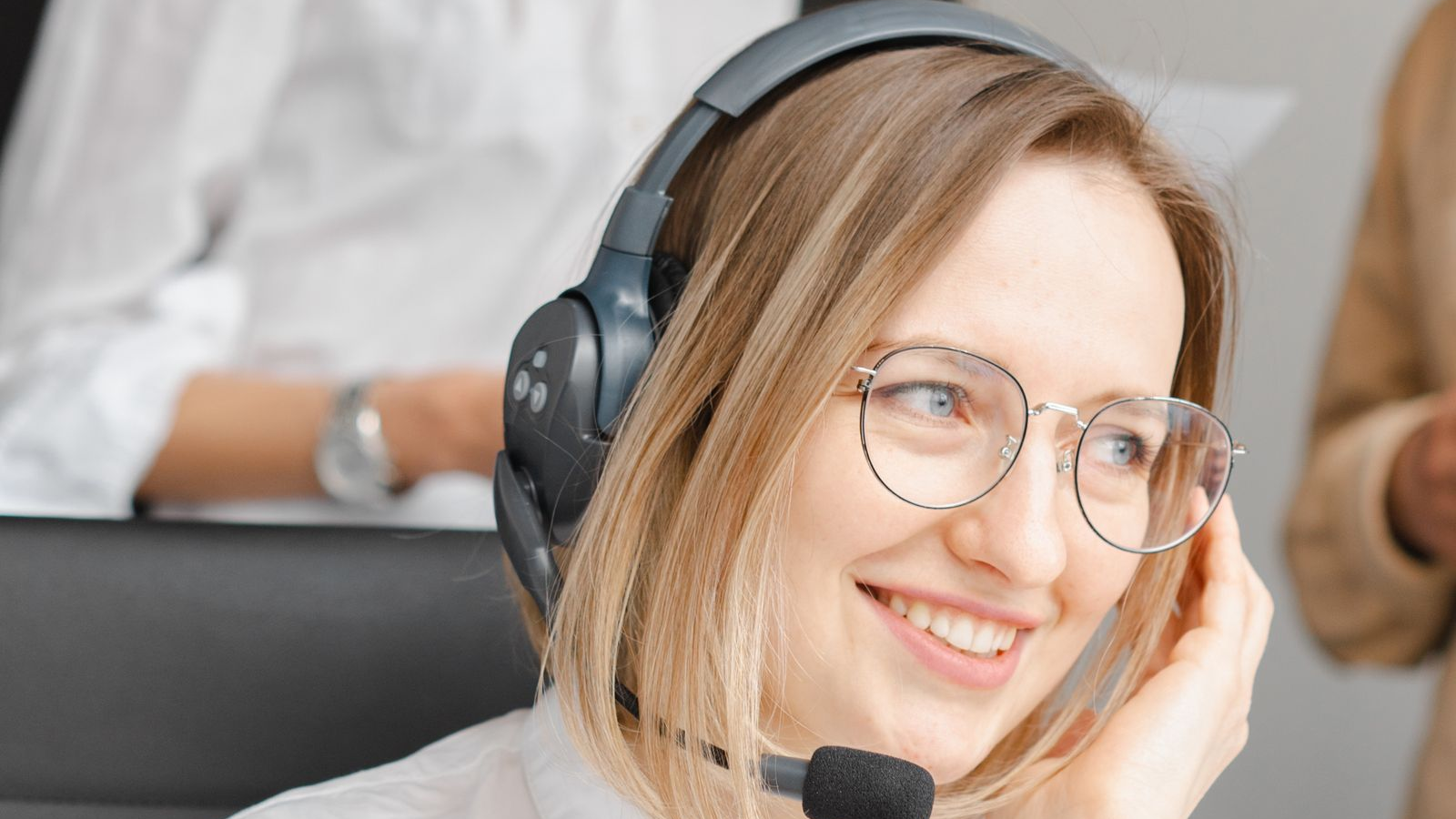 Smiling sales representative with glasses wearing a black headset at a laptop on a shared dial-floor style office, with colleagues visible in the blurred background.