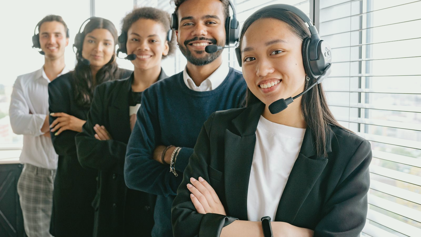 Mid-market sales team wearing headsets in a bright office, human faces visible in a diagonal line; real BD or sales-floor call environment with natural window light.