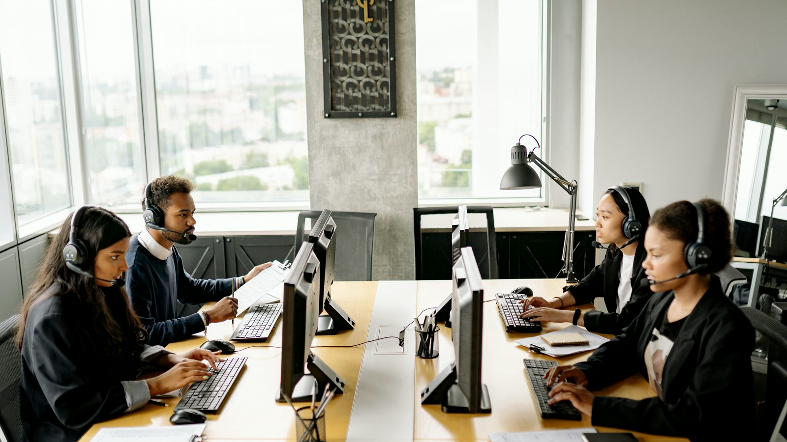 Four professionals with headsets working at a shared call desk with monitors and clipboards in a bright office, freight-brokerage-adjacent phone floor.