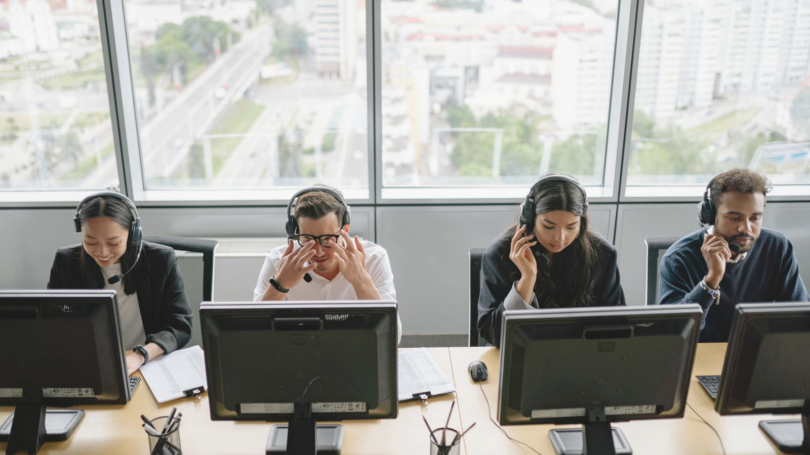 Diverse call-desk team with headsets at a shared office desk; one colleague shows scramble stress while others stay on live calls, freight-brokerage-adjacent ops floor.
