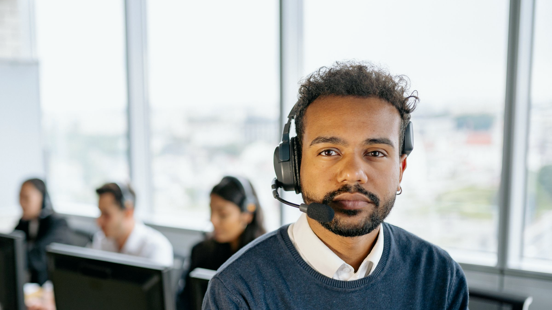Composed freight-brokerage-adjacent rate-desk broker with headset on a live call floor; colleagues on headsets in the background, calm focus after putting a number on the table.