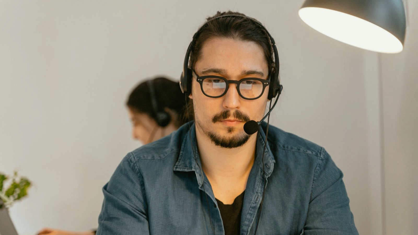 Freight-brokerage-adjacent ops professional with a headset at a desk laptop, focused expression under warm desk light, colleague on headset soft in the background.