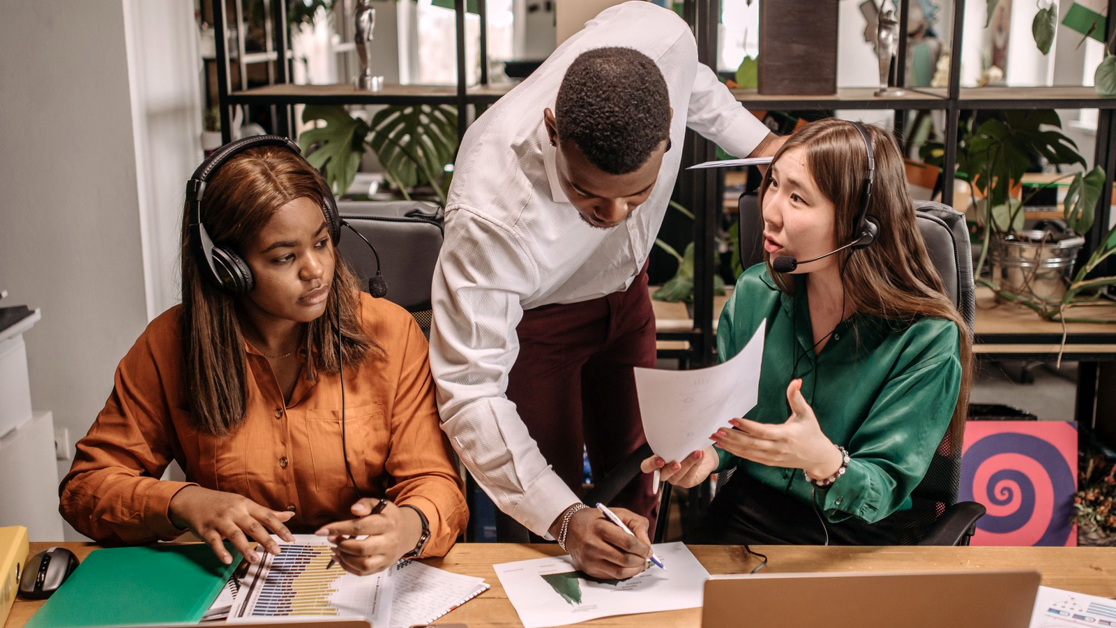 Three sales-floor teammates with headsets reviewing charts at a shared desk, human faces on a mid-market SDR/BDR-style floor where motion scoreboards still look green.