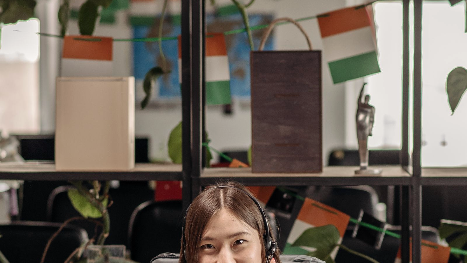 Woman with headset at a wooden desk in a plant-filled office, mid-call, human face on a real discovery-desk environment for individual SDR/BDR/AE presence.