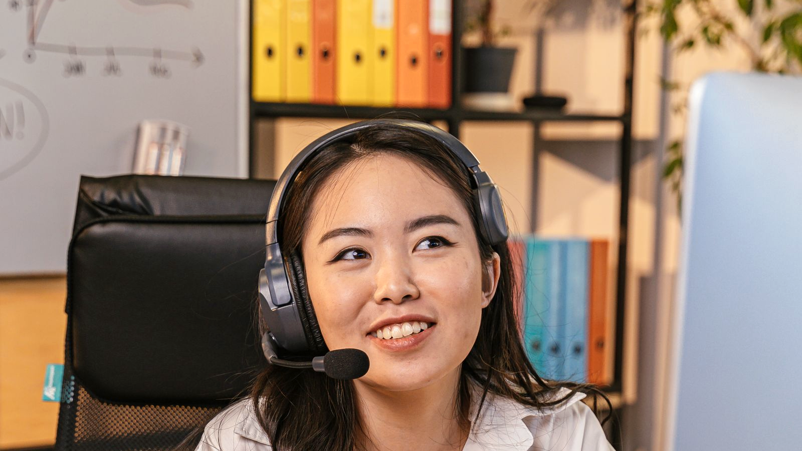 Woman with headset at an office desk with notebook and monitor, listening mid-call, human face on a real AE/SDR dialer desk environment.