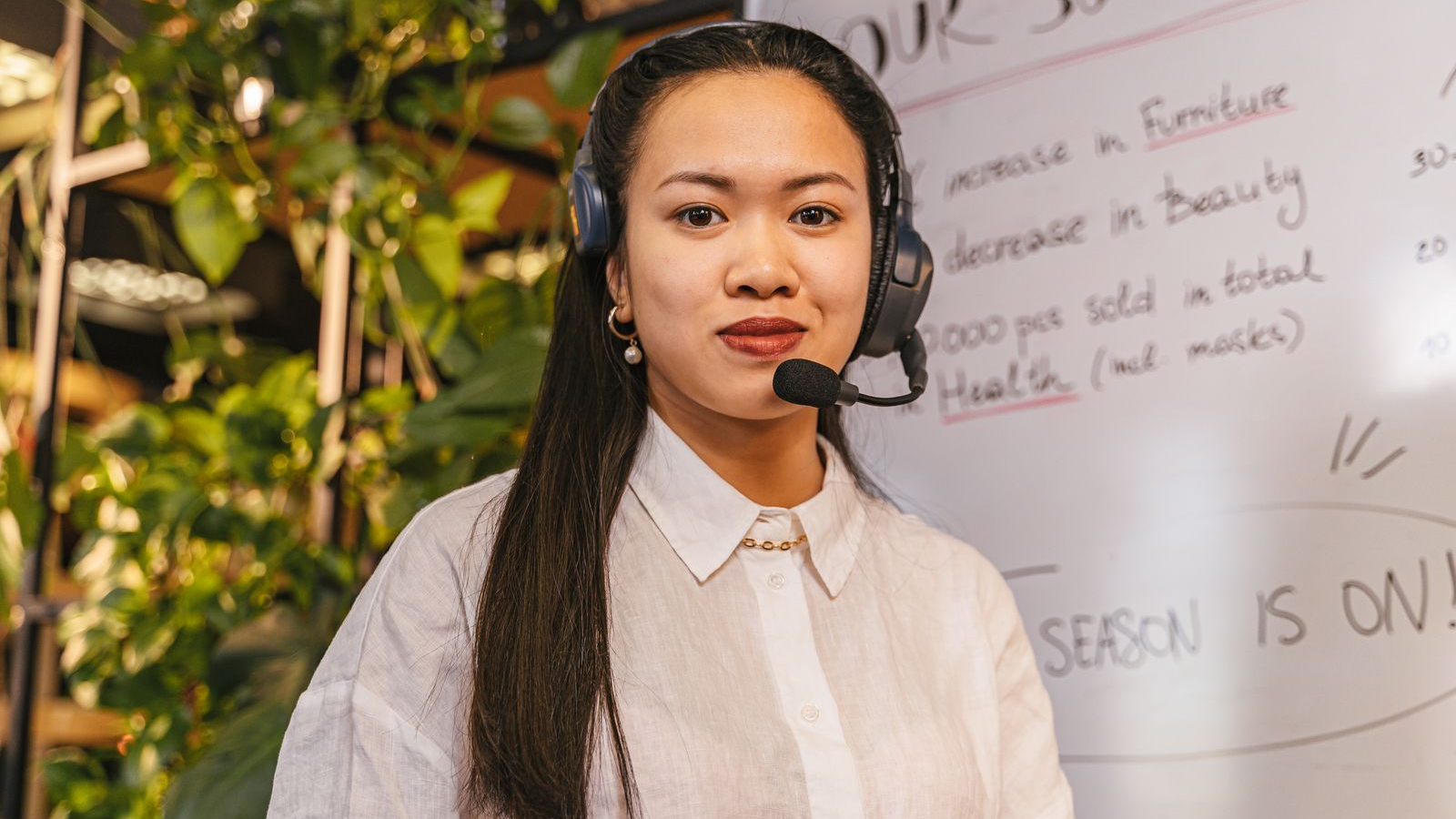 Woman wearing a headset at a desk in a bright office, with a handwritten whiteboard and plants behind her.