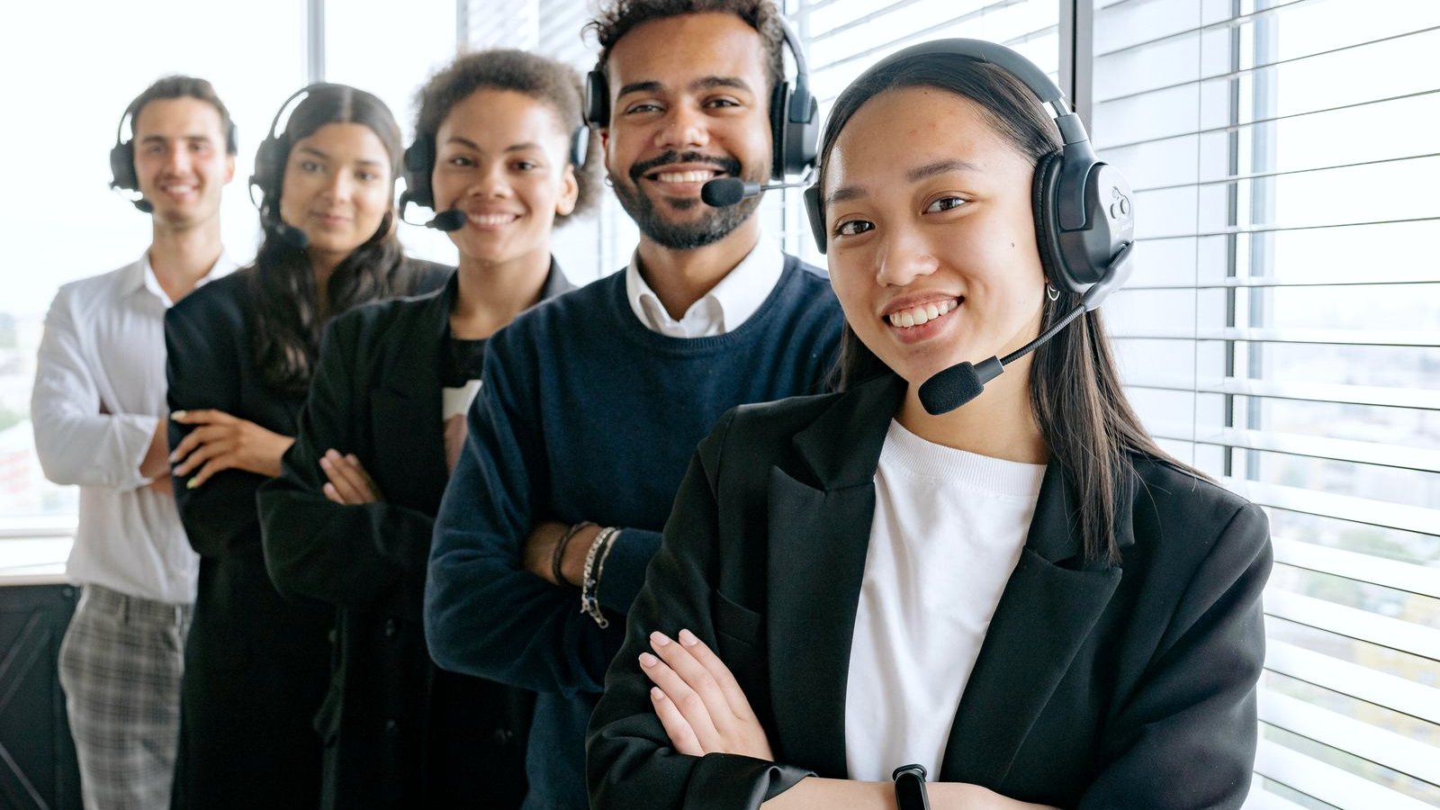 Diverse call-center and BD team standing in a bright office, each wearing a black headset with boom mic, smiling toward the camera.