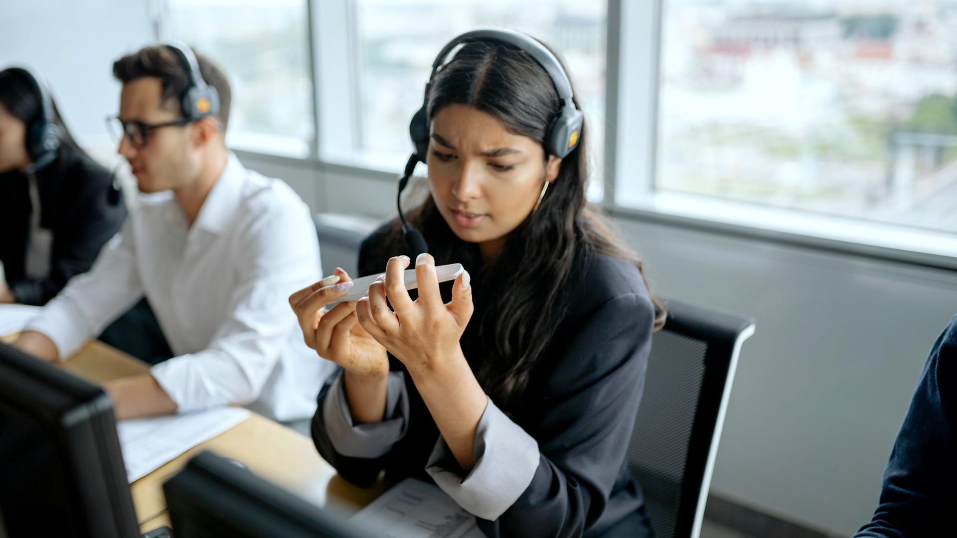 Woman wearing a professional headset with boom mic at an open-office phone desk, looking at her phone, with colleagues also wearing headsets working behind her near large windows.