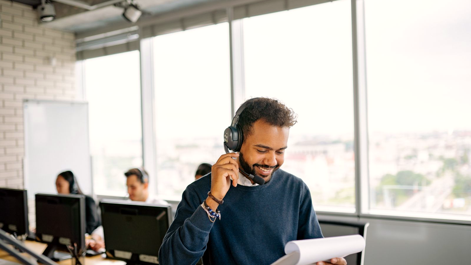 Staffing or recruiting BD professional smiling while on a headset call at a bright open desk, clipboard in hand, colleagues on headsets visible behind; human faces in a real call-heavy sales floor.