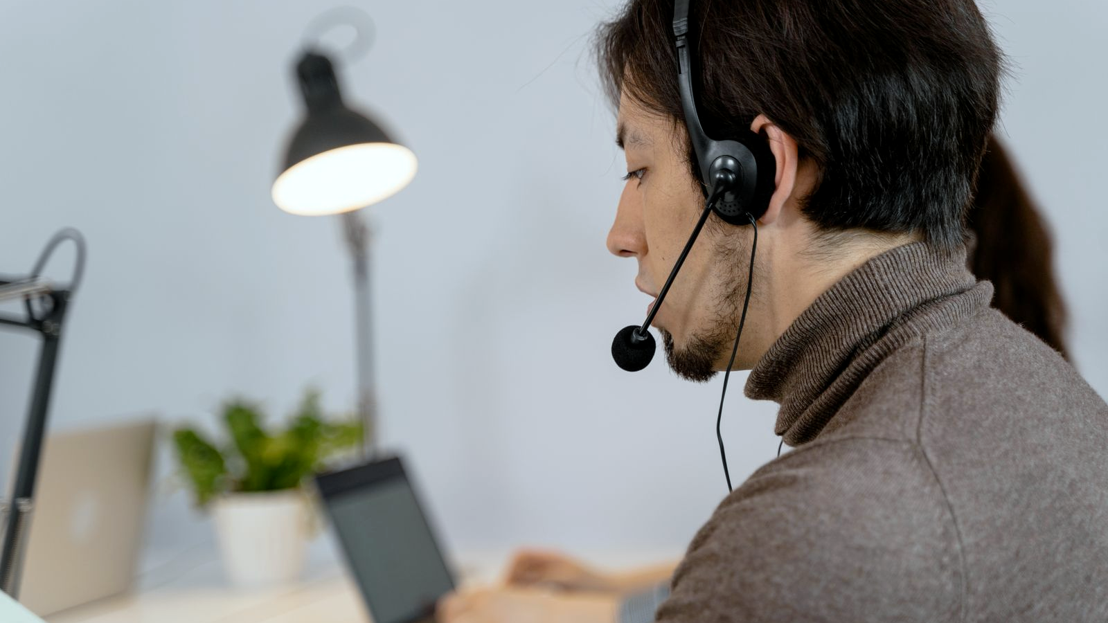 Professional wearing a headset at an open-office desk, looking focused after a call, with a colleague typing on a laptop behind; staffing BD / recruiting sales floor.