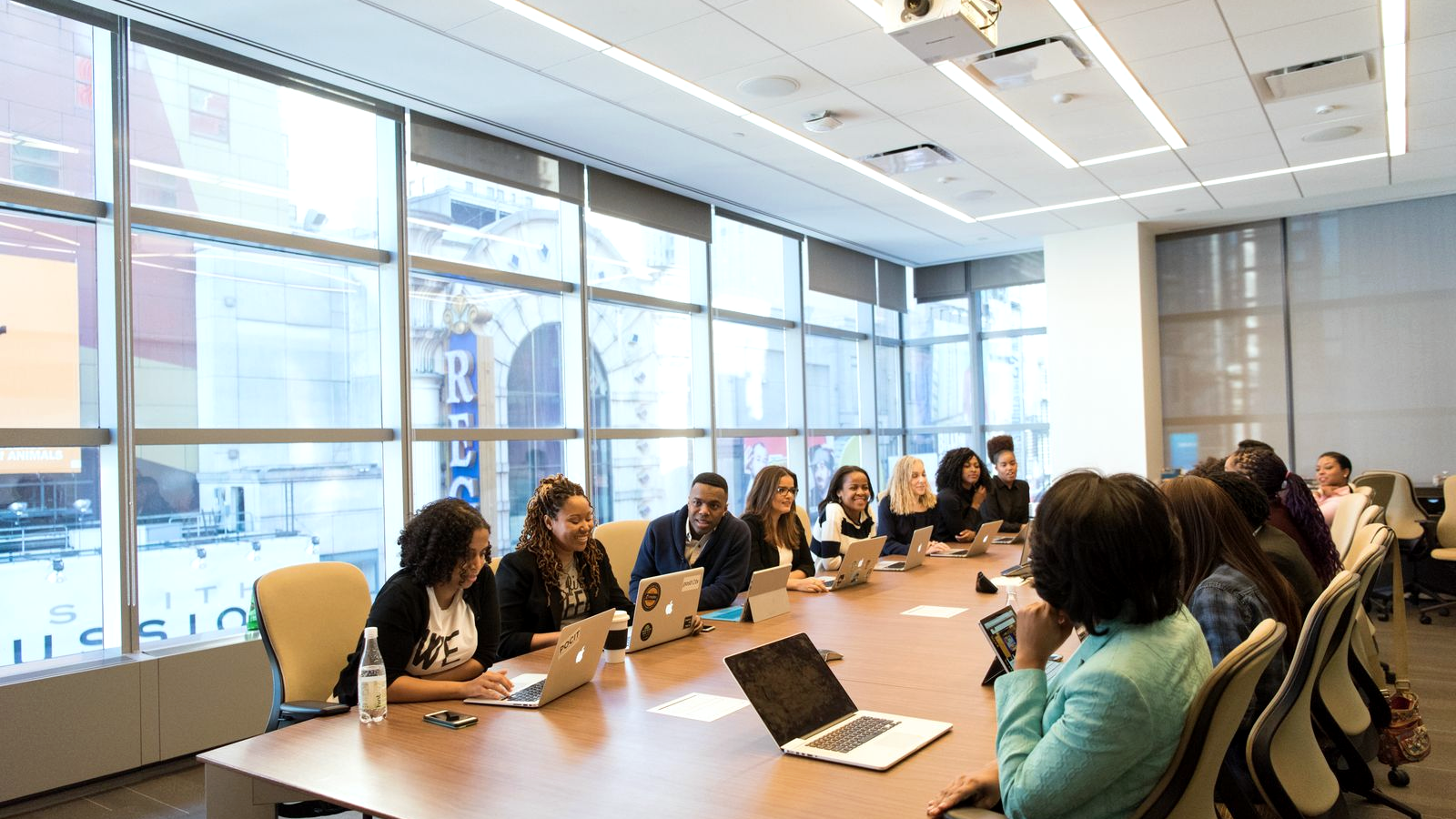 Real boardroom conference table with human faces; presenter in a teal jacket addressing colleagues around open laptops in a bright meeting room.