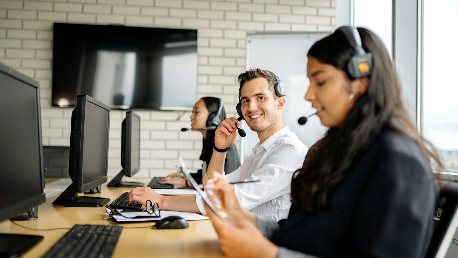 Three call-heavy sellers at a shared desk with headsets and monitors in a bright office; central professional smiling while adjusting a boom mic, human faces in a real sales-floor talk environment.